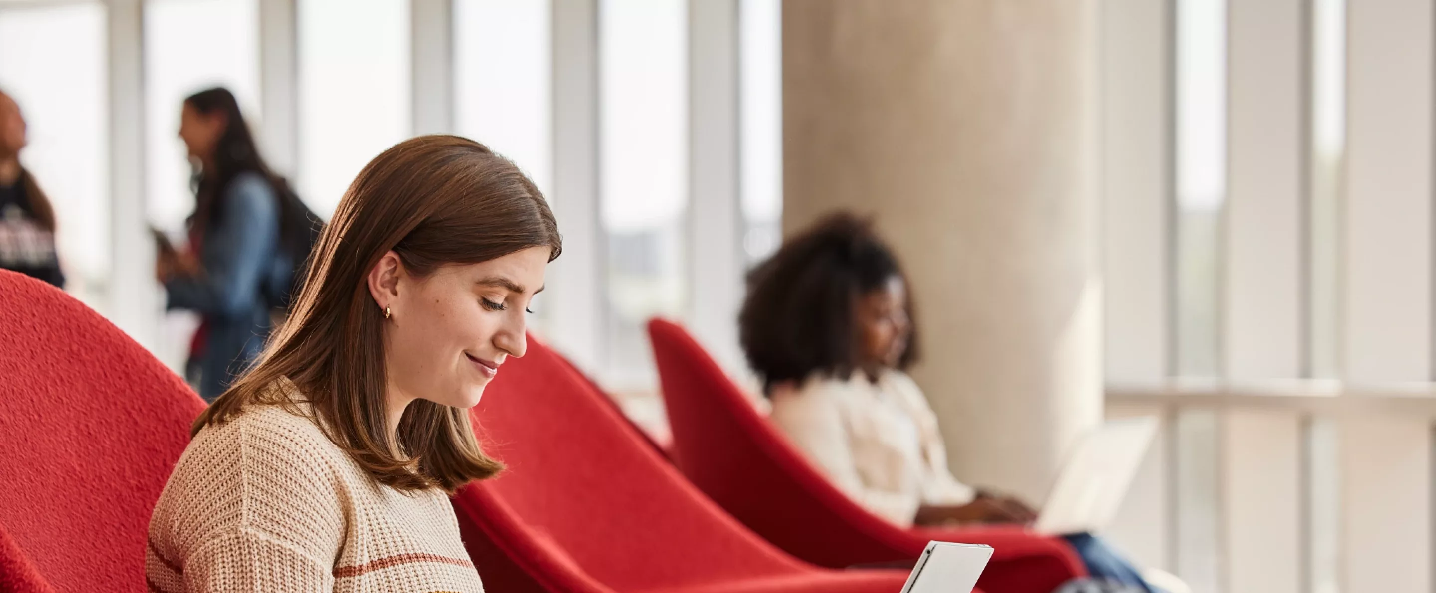 Lexi Raikes sitting on the second floor of the Belknap Academic Building, in a red chair, typing happily on her laptop
