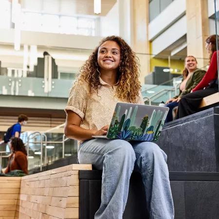 Student recognizes another student walk into the Belknap Academic Building as she is typing on her laptop and sitting on the wooden platform benches at the entrance of the BAB.